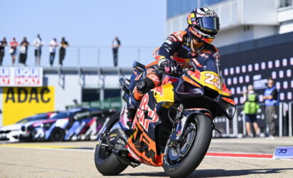 Moto racer on orange-red KTM bike numbered 25 doing a sharp turn on a race track; Red Bull branding visible, spectators in background.