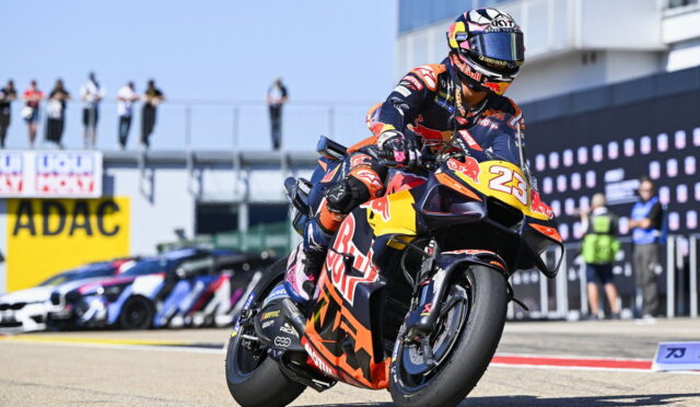 Moto racer on orange-red KTM bike numbered 25 doing a sharp turn on a race track; Red Bull branding visible, spectators in background.