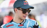 Rider in a blue racing suit with sponsor logos, wearing a Red Bull cap and sunglasses, looking to the side.