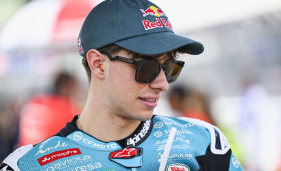 Rider in a blue racing suit with sponsor logos, wearing a Red Bull cap and sunglasses, looking to the side.