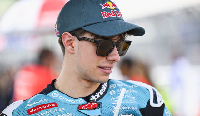 Rider in a blue racing suit with sponsor logos, wearing a Red Bull cap and sunglasses, looking to the side.