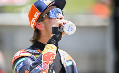 Motocross rider in orange-blue gear drinks water from a bottle during a break.