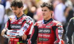 Two motorcycle racers in red Lenovo racing suits stand with arms crossed, spectators blurred in the background.