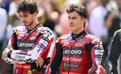 Two motorcycle racers in red Lenovo racing suits stand with arms crossed, spectators blurred in the background.