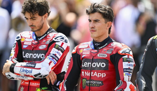 Two motorcycle racers in red Lenovo racing suits stand with arms crossed, spectators blurred in the background.