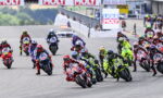 Group of motorcycle racers leaned into a tight turn on a racetrack, colorful bikes and gear, with banners in the background.