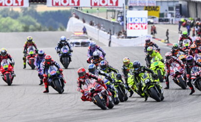 Group of motorcycle racers leaned into a tight turn on a racetrack, colorful bikes and gear, with banners in the background.