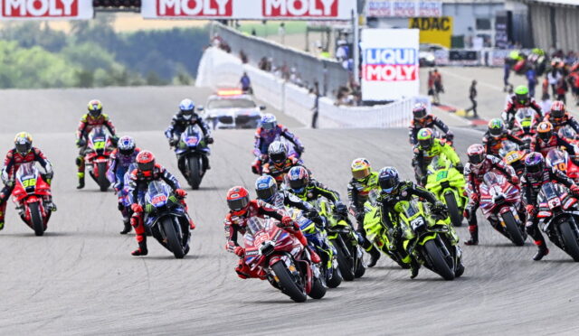 Group of motorcycle racers leaned into a tight turn on a racetrack, colorful bikes and gear, with banners in the background.