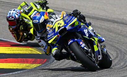 Two motorcycle racers lean into a right-hand curve on a race track; the lead rider rides a blue-yellow bike with number 73 and sponsor decals, closely followed by a neon yellow rider behind.