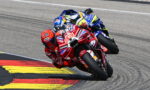 Two motorcycle racers lean into a sharp corner, red Ducati leading a blue-yellow bike close behind on a race track with red-yellow curbing.