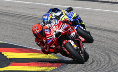 Two motorcycle racers lean into a sharp corner, red Ducati leading a blue-yellow bike close behind on a race track with red-yellow curbing.