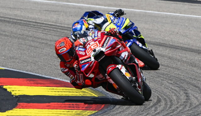 Two motorcycle racers lean into a sharp corner, red Ducati leading a blue-yellow bike close behind on a race track with red-yellow curbing.