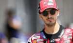 Male motorcyclist in a red cap with sponsor logos and sunglasses, head-and-shoulders portrait in racing gear.