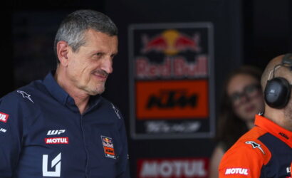 Two men in racing team uniforms stand in a garage area; one in navy with sponsor logos smiles, the other in orange with a headset nearby, Red Bull/ KTM signage in the background.