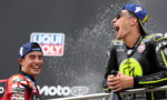 Two motorcycle racers on a podium spraying champagne, laughing, in racing suits with sponsor logos.