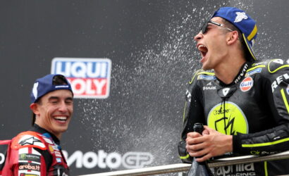 Two motorcycle racers on a podium spraying champagne, laughing, in racing suits with sponsor logos.