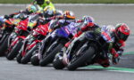 Group of motorcycle racers leaning into a tight turn on a race track in a close pack, tires screeching on asphalt.
