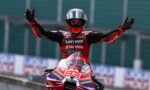 MotoGP rider in a red Lenovo racing suit raises arms in victory on the track