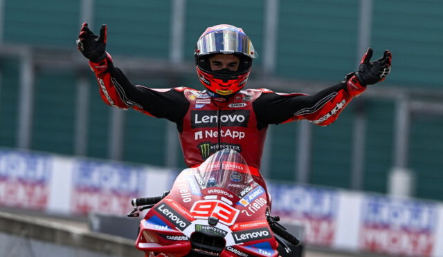 MotoGP rider in a red Lenovo racing suit raises arms in victory on the track