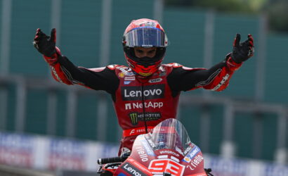 Moto racer in red suit raises arms in victory atop a red bike with number 93, Lenovo and NetApp sponsorships visible.