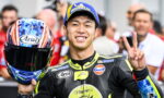 Moto racer in black suit smiling and making a peace sign while holding a colorful helmet with Arai logo, crowd behind him.