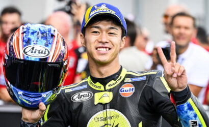Moto racer in black suit smiling and making a peace sign while holding a colorful helmet with Arai logo, crowd behind him.
