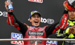 Moto racer on a podium celebrating with arms raised, holding a helmet in one hand and a spray can in the other.