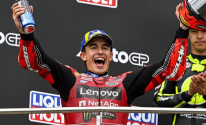 Moto racer on a podium celebrating with arms raised, holding a helmet in one hand and a spray can in the other.
