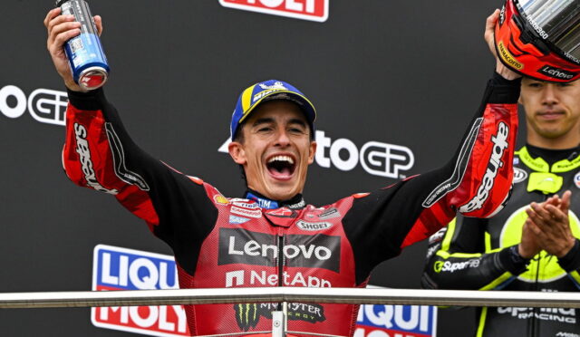 Moto racer on a podium celebrating with arms raised, holding a helmet in one hand and a spray can in the other.