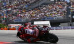Motorcycle racer in red leans into a corner on a race track, with a packed grandstand in the background.
