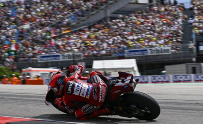 Motorcycle racer in red leans into a corner on a race track, with a packed grandstand in the background.