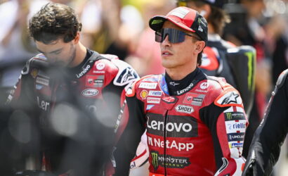 MotoGP rider in red Lenovo racing suit with sponsor logos, wearing cap and visor sunglasses, in a crowded pit area.