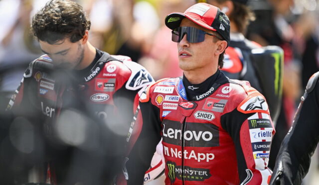 MotoGP rider in red Lenovo racing suit with sponsor logos, wearing cap and visor sunglasses, in a crowded pit area.