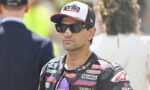 Racer wearing a white and black cap with Aprilia Racing logo and dark sunglasses, in a branded racing suit.