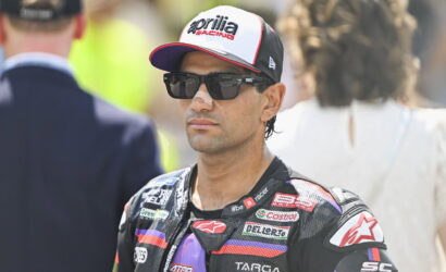 Racer wearing a white and black cap with Aprilia Racing logo and dark sunglasses, in a branded racing suit.