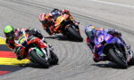 Three professional motorcycle racers lean into a sharp turn on a race track, colorful bikes close together.",