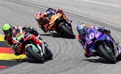 Three professional motorcycle racers lean into a sharp turn on a race track, colorful bikes close together.",