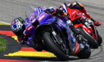 Three riders lean into a turn on a race track, foreground purple Yamaha bike leading a pack behind on the red-yellow curbed circuit in sunlight and spray.