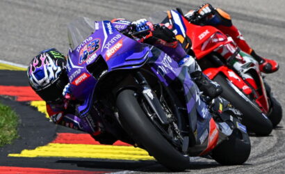 Three riders lean into a turn on a race track, foreground purple Yamaha bike leading a pack behind on the red-yellow curbed circuit in sunlight and spray.