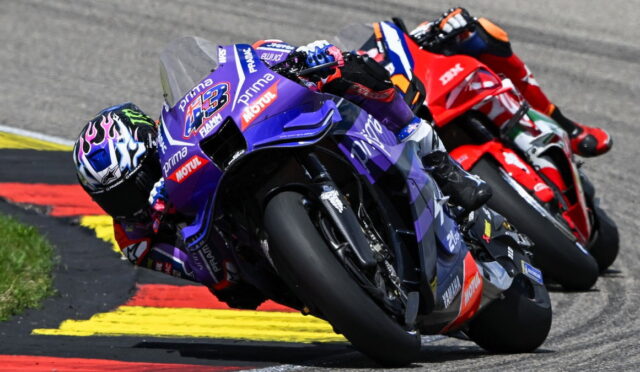 Three riders lean into a turn on a race track, foreground purple Yamaha bike leading a pack behind on the red-yellow curbed circuit in sunlight and spray.