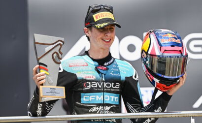 Moto rider on a podium holding a trophy in his left hand and a Red Bull helmet in his right.
