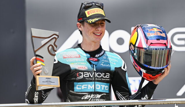 Moto rider on a podium holding a trophy in his left hand and a Red Bull helmet in his right.