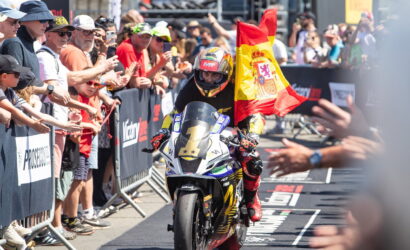 Moto racer on a race bike crosses the finish line as a crowd cheers and a Spanish flag is waved in the background at a busy race event, with barriers and spectators on both sides.
