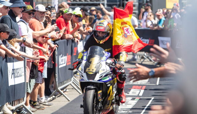 Moto racer on a race bike crosses the finish line as a crowd cheers and a Spanish flag is waved in the background at a busy race event, with barriers and spectators on both sides.
