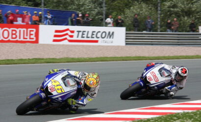 Two motorcycle racers lean into a sharp turn on a race track, with blue Yamaha bikes and colorful helmets as spectators watch from behind barriers in the background.