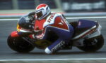 Motorcycle racer in red, white, and blue leathers leaning into a corner on a sportbike during a race.