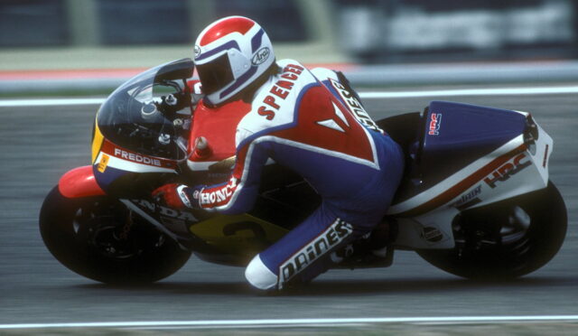 Motorcycle racer in red, white, and blue leathers leaning into a corner on a sportbike during a race.