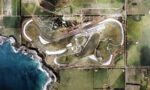 Aerial view of a coastal racing circuit with a winding track, paddocks, and surrounding fields beside the ocean.