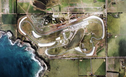 Aerial view of a coastal racing circuit with a winding track, paddocks, and surrounding fields beside the ocean.