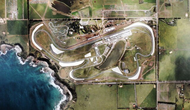 Aerial view of a coastal racing circuit with a winding track, paddocks, and surrounding fields beside the ocean.